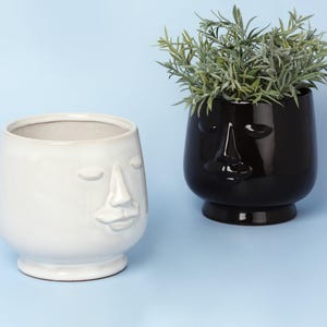 Op de afbeelding: Twee keramische gezichtspotten, één wit en één zwart, staan op een lichtblauwe ondergrond. De witte pot heeft een matte afwerking, de zwarte is glanzend en bevat groen gebladerte. Beide potten hebben een gezichtsontwerp.