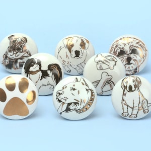 Könnte beinhalten: Satz von acht weißen Keramik-Knöpfen mit goldenen Akzenten, die verschiedene Hunderassen und ein Pfotenabdruck-Design zeigen.