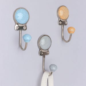 May include: Three decorative wall hooks with a crackle glaze design. The hooks have a silver finish and are topped with round ceramic knobs in blue, grey, and orange. A white strap hangs from one of the hooks.
