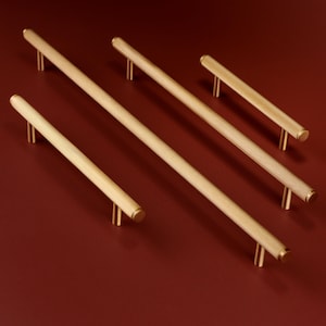 Könnte beinhalten: Fünf goldfarbene Schrankgriffe mit geriffeltem Design. Die Griffe sind diagonal auf einer tiefroten Oberfläche angeordnet, wodurch ihre zylindrische Form und minimalistische Ästhetik hervorgehoben werden. Geeignet für Schubladen und Schränke.