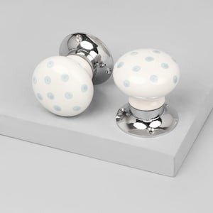 May include: Two white ceramic door knobs with a light blue polka dot pattern. The knobs have a rounded shape and are mounted on shiny chrome bases. The door hardware is displayed on a light grey surface.