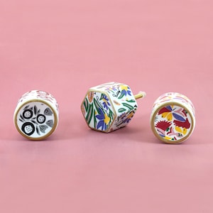 Puede incluir: Tres pomos decorativos de cerámica con diseños florales. Los pomos tienen una base blanca con flores coloridas, ribete dorado y fondo rosa. Un pomo es hexagonal y los otros dos son cilíndricos.