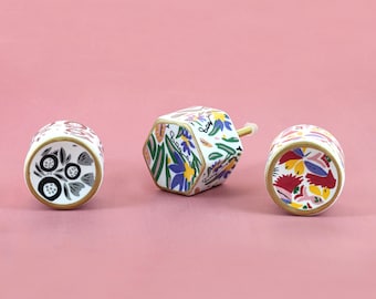 May include: Three decorative ceramic knobs with floral designs. The knobs have a white base with colorful flowers, gold trim, and a pink background. One knob is hexagonal, and the other two are cylindrical.