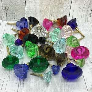 May include: A selection of colourful glass cabinet knobs in jewel tones such as blue, green, pink, and clear. Each knob features a faceted design and a brass screw. The knobs are arranged on a light wood-grain surface.