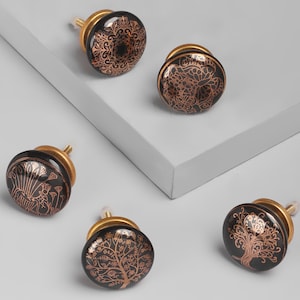 Peut inclure: Ensemble de cinq boutons de cabinet en céramique noire avec des accents dorés. Chaque bouton présente un motif complexe différent, y compris un arbre, un motif floral et un motif géométrique.