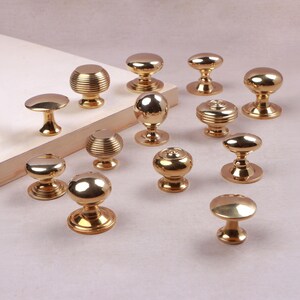 May include: A collection of twelve polished gold-tone metal cabinet knobs in various shapes, including smooth spherical, flat-topped, and horizontally ribbed designs, each featuring a circular base for mounting to furniture or cabinetry doors.