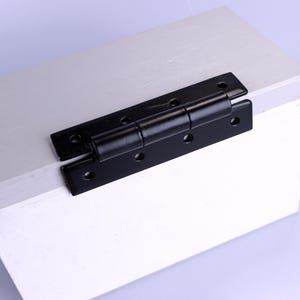 May include: A black metal hinge with multiple screw holes is mounted on a white wooden surface. The hinge is in the open position, showing its two rectangular leaves connected by a cylindrical barrel.