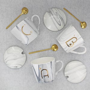 May include: A set of white ceramic mugs with a marble design, gold spoons, and matching lids. Each mug has a gold letter and symbol. The mugs are arranged on a light gray surface.