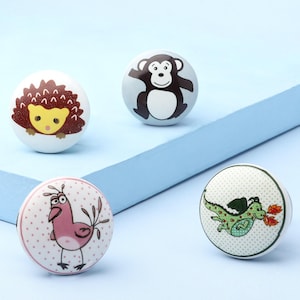 May include: Four decorative ceramic cabinet knobs with animal designs. The knobs feature a hedgehog, monkey, bird, and dragon. The hedgehog has brown and yellow details, the monkey is brown, the bird is pink, and the dragon is green. Each knob is round and white.