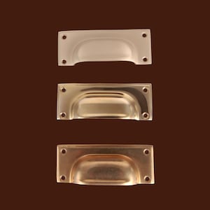 Peut inclure: Trois poignées de tiroir rectangulaires en métal disposées verticalement. Chaque poignée présente une prise centrale encastrée et quatre trous de vis circulaires, un à chaque coin. La poignée supérieure est argent mat, celle du milieu or poli, et celle du bas cuivre.