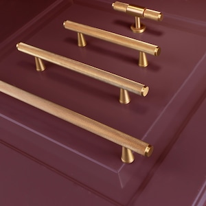 Peut inclure: Quincaillerie de placard dorée, comprenant plusieurs barres de tirage et un bouton en T, disposée sur une surface violet foncé. Les poignées ont un motif texturé et pointillé. La quincaillerie est probablement pour les armoires de cuisine ou de salle de bain.