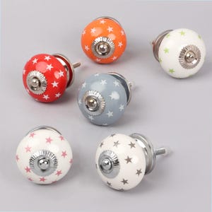 Può includere: Una collezione di sei pomelli decorativi per mobili. Ogni pomello è di un colore diverso, tra cui rosso, arancione, blu, bianco e verde. Ogni pomello è decorato con un motivo a stelle e ha una base color argento.