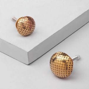 May include: Two gold-toned cabinet knobs with a woven pattern. The knobs are round and have a screw on the back.