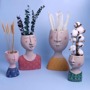 Op de afbeelding: Vier decoratieve gezichtsvormige plantenbakken in verschillende maten en kleuren. De plantenbakken zijn geschilderd in tinten roze, grijs en blauw, met details zoals blozen en kleding met patronen. Elke plantenbak bevat verschillende soorten gedroogde planten en katoen.