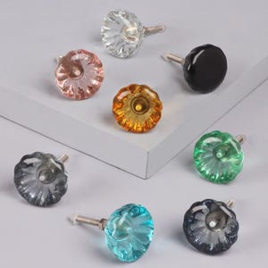 May include: A selection of decorative glass cabinet knobs in assorted colours: clear, pink, amber, black, green, blue, and grey. Each knob features a fluted, flower-like design and a silver-toned screw.
