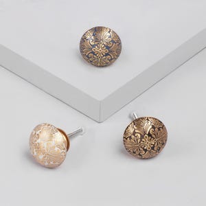 Pode incluir: Três puxadores decorativos para armários com padrões florais. Um é branco com detalhes dourados, outro azul marinho com dourado e o terceiro dourado com fundo escuro. Cada puxador tem um parafuso prateado.