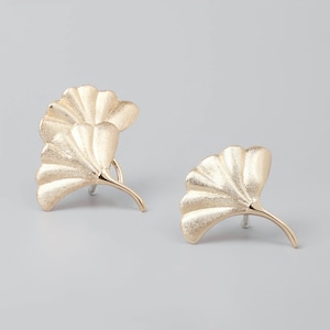 May include: Two gold-colored ginkgo leaf earrings. The earrings have a textured surface and a curved stem. The leaves are detailed with multiple veins. The earrings are set against a light gray background.