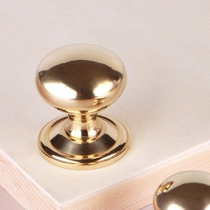 May include: Two polished brass cabinet knobs with a smooth, rounded mushroom-shaped head and a circular base plate. The metallic finish is reflective and gold-toned, featuring a classic silhouette with a tapered neck connecting the top to the base.