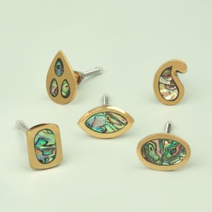 Può includere: Una collezione di cinque pomelli decorativi per mobili di varie forme. Ogni pomello presenta una cornice in metallo dorato con un intarsio iridescente di conchiglia di abalone. Le forme includono una goccia, un occhio, un paisley, un quadrato e un ovale.