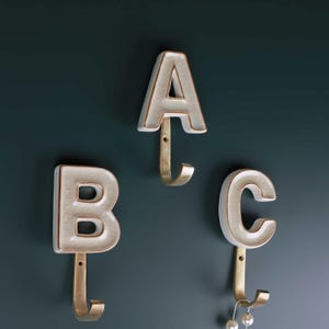 Ganchos de pared con letras de latón con acabado antiguo y cerámica pintada a mano con monograma personalizado de G Decor, con esmalte craquelado crema y letras del alfabeto.