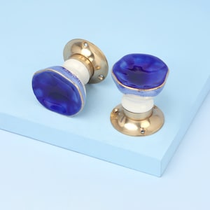Può includere: Due pomelli per porte in ceramica blu e bianca con dettagli dorati. I pomelli sono su uno sfondo blu chiaro.