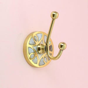 May include: A brass wall hook with a decorative circular base featuring a white and gold pattern. The hook has a curved design with two rounded ends. The hook is mounted on a pink wall.