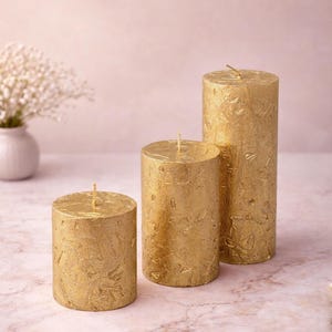 May include: Three gold pillar candles of varying heights. The candles have a textured surface and a wick at the top. The candles are cylindrical and are arranged on a light pink surface.