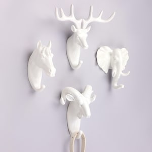 May include: Four white animal head wall hooks: a horse, a deer, an elephant, and a ram. Each hook is sculpted in a realistic style and mounted on a light purple wall. The deer has large antlers, and the elephant has a curled trunk.