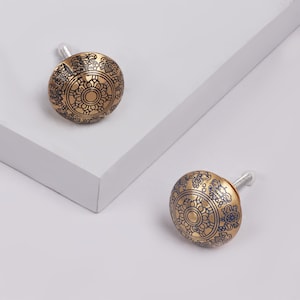 May include: Two gold-coloured cabinet knobs with intricate blue and black designs. The knobs are round and have a slightly domed shape.