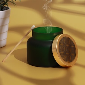 May include: A dark green candle jar with a gold lid featuring an ornate design. A lit match rests against the jar, and smoke rises from the opening. The jar sits on a yellow surface, suggesting a home decor item.