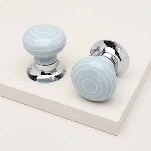 May include: Two round door knobs featuring a light blue ceramic finish with concentric white circular patterns. Each knob is attached to a polished chrome circular base plate with visible screw holes for mounting to a door or cabinet surface.