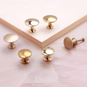 May include: A collection of six gold-toned metal cabinet knobs with a smooth, rounded top and a tapered base. Some knobs feature a polished, reflective finish, while others have a matte, satin appearance. One knob is shown from the side.