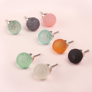 Può includere: Una collezione di pomelli per armadietti in vetro sfaccettato colorato in varie tonalità tra cui rosa, verde, arancione, grigio e nero. Ogni pomello ha una vite color argento per il fissaggio. I pomelli sono disposti su una superficie rosa chiaro.