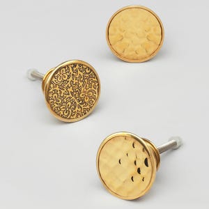 Puede incluir: Tres tiradores dorados para armarios con diseños decorativos. Dos tienen una textura martillada, el tercero un patrón floral. Cada tirador tiene un tornillo plateado. Ideal para muebles. Diámetro aproximado de 3 cm.