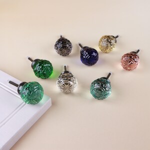 May include: A collection of eight round, faceted glass cabinet knobs in various colors including green, clear, blue, yellow, and pink. Each knob features a textured, geometric pattern and a metal screw post for installation.