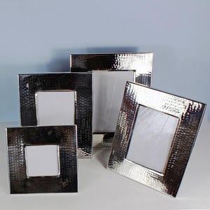 May include: A collection of four silver-colored picture frames with a hammered texture. The frames are square with a white interior and vary in size, arranged on a white surface against a light blue background. The frames are made of metal.