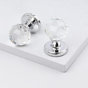May include: Two crystal door knobs with faceted spherical handles and polished chrome bases. One knob is lying on its side, while the other is upright. The knobs are on a white surface.