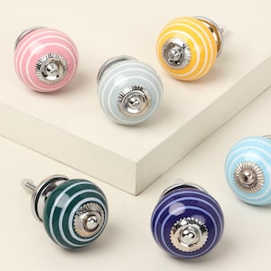 Puede incluir: Seis coloridos tiradores de cerámica para armarios. Cada tirador es de un color diferente: rosa, amarillo, azul, verde, gris y morado. Cada tirador tiene rayas blancas y una base de metal plateado. Son herrajes decorativos.