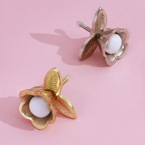 Peut inclure: Deux boutons de meuble décoratifs en forme de fleur, l'un doré et l'autre argenté. Chaque bouton a un centre blanc et sphérique. Les boutons sont posés sur un fond rose clair.