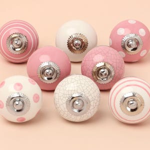 May include: Eight ceramic cabinet knobs in a variety of designs. The knobs feature pink and white stripes, polka dots, and crackle patterns. Each knob has a silver-toned metal base and a round shape, perfect for drawers or cupboards.
