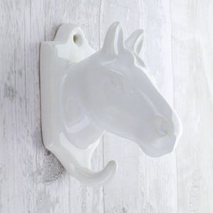 Può includere: Un gancio da parete a forma di testa di cavallo in ceramica bianca. Il gancio ha la forma di una testa di cavallo con un gancio curvo che si estende dal basso. Il gancio ha un foro nella parte superiore per appenderlo.