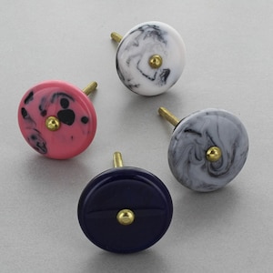 May include: Four decorative cabinet knobs in various colors. One is pink with black speckles, another is navy blue, and two have a marbled effect in white and gray. Each knob has a gold-colored screw.