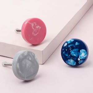 Può includere: Tre pomelli decorativi per armadi in rosa, grigio e blu. Ogni pomello è rotondo con un design marmorizzato e una vite argentata. Il pomello rosa ha venature bianche, il grigio ha venature bianche e il blu ha venature bianche e azzurre.