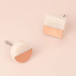 Può includere: Due pomelli decorativi per mobili. Uno è rotondo, l'altro quadrato. Entrambi hanno una parte superiore in marmo bianco e una metà inferiore color rame. I pomelli hanno uno stelo argentato per il montaggio. Lo sfondo è rosa chiaro.