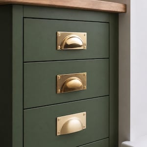 Puede incluir: Un mueble de madera de color verde oliva oscuro con tres cajones apilados. Cada cajón cuenta con un tirador de concha de latón pulido, fijado con cuatro tornillos pequeños en las esquinas de la placa metálica rectangular de cada tirador.