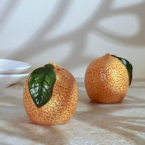 May include: Two orange-shaped ceramic salt and pepper shakers. Each shaker has a textured orange peel design, a green leaf, and small holes on top. The shakers are on a light-colored surface.