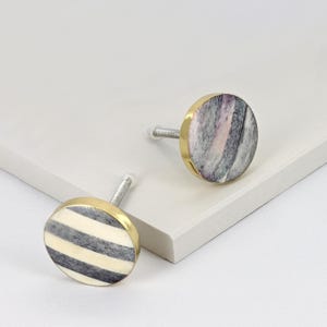 Op de afbeelding: Twee decoratieve kastknoppen. Eén knop heeft een cirkelvormig ontwerp met afwisselende zwarte en crèmekleurige strepen. De andere knop heeft een gemarmerd uiterlijk met tinten grijs, roze en wit. Beide knoppen hebben een goudkleurige rand.