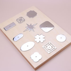 May include: A collection of eleven polished silver-toned metal shapes with central holes, including stars, flowers, rectangles, ovals, and squares, arranged on a light-colored wooden board. Each piece features a smooth, reflective surface.