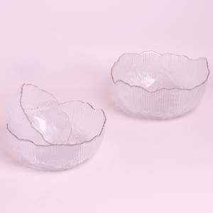 May include: Three clear glass bowls with a textured, ribbed design and gold-coloured rims. The bowls are arranged on a light pink background. The bowls have an organic, wavy shape.