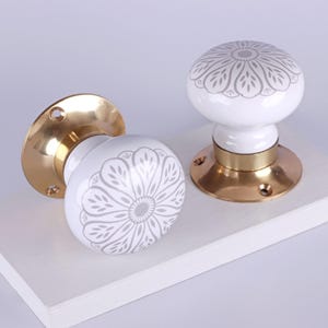 May include: Two white ceramic door knobs with a gray floral design. The knobs have a brass base and are displayed on a white surface. The knobs are a decorative hardware item.